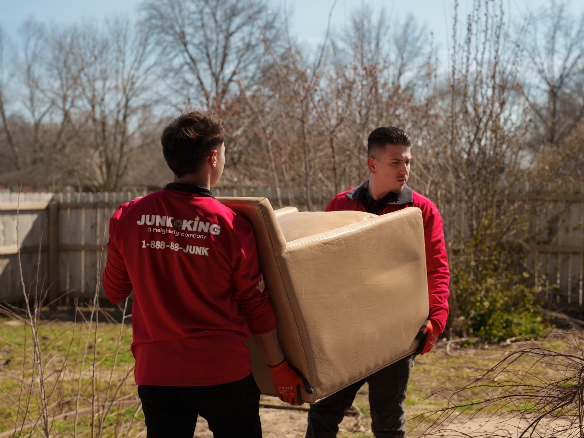 Two Junk King crew members in branded red uniforms carrying an upholstered armchair through a customer's backyard